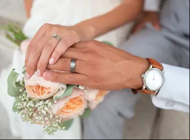 Imagen que captura las manos unidas de una pareja con anillos de matrimonio, simbolizando su unión legal y afectiva, con un delicado ramo de flores de boda proporcionando un hermoso fondo desenfocado, representando el compromiso y la ceremonia de matrimonio en la cultura contemporánea.